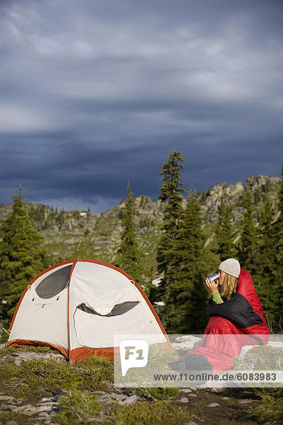 Adult woman drinking coffee in a sleeping bag near her tent while backpack camping.