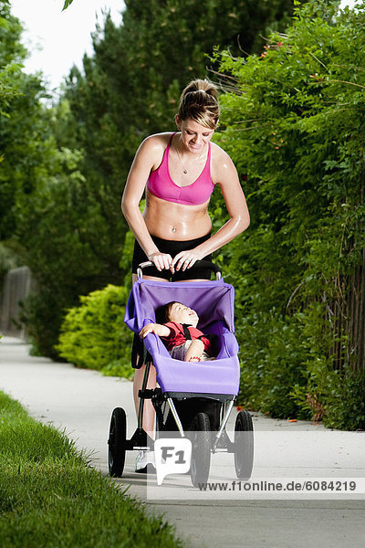 An edgy-looking  athletic young woman looks down at a smiling baby in his jogging stroller during a run on a suburban sidewalk.