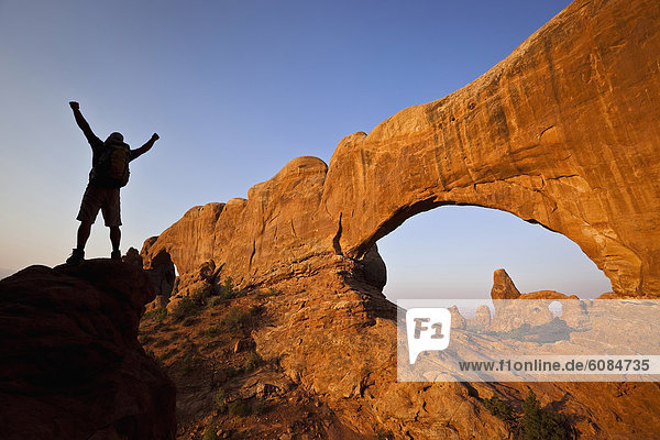 A silhouetted hiker raises his arms beside an arch in Arches National Park  Utah.