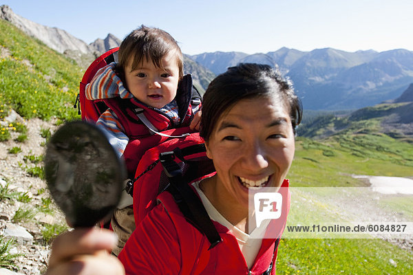 A mother looks at her 14 month old son with a hand held mirror while she carries him in a backpack over Trail Rider Pass amongst a wildflower filled alpine meadow in the Maroon Bells in Snowmass Wilderness just outside of Aspen.