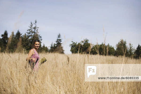 A young woman  wearing a purple shirt  carries a green yoga mat through a field of golden grasses.