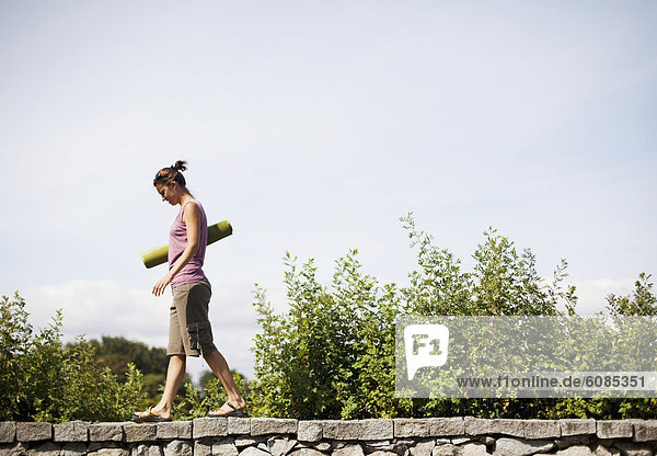 A young woman walks on a stone ledge holding a green yoga mat.
