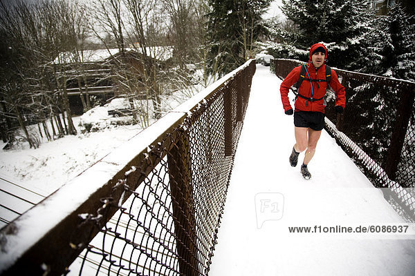 A male runner with a backpack runs across a snow-covered bridge while wearing shorts in the winter.