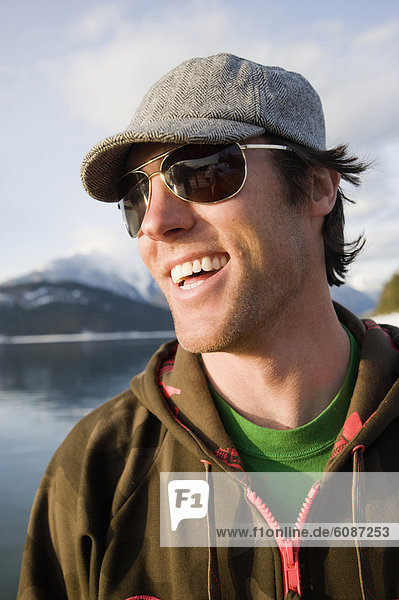 A young man in sunglasses and a hat laughs on the shores of a river.