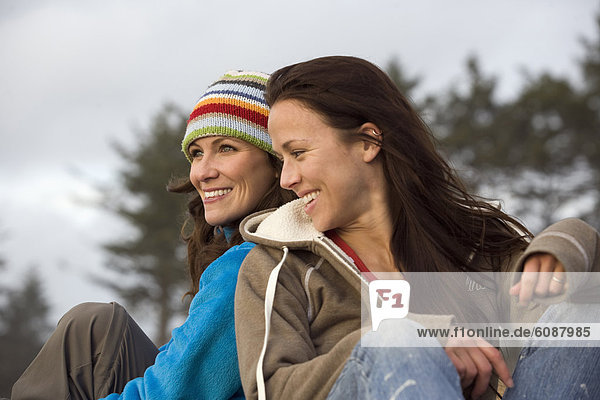 Two young women sit back to back in a open forest setting  smiling and laughing.