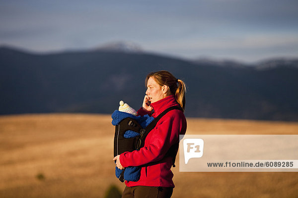 Mother on cell phone and newborn baby in front carrier while hiking through grassy hills  Water Works  Missoula  Montana.