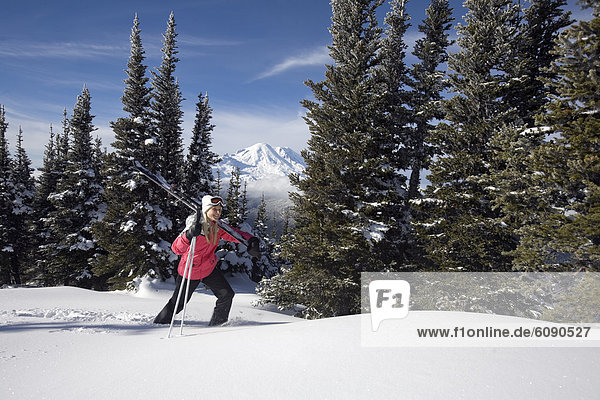 A woman carrying skis and poles hikes in snow with Mt. Rainier views in the background.