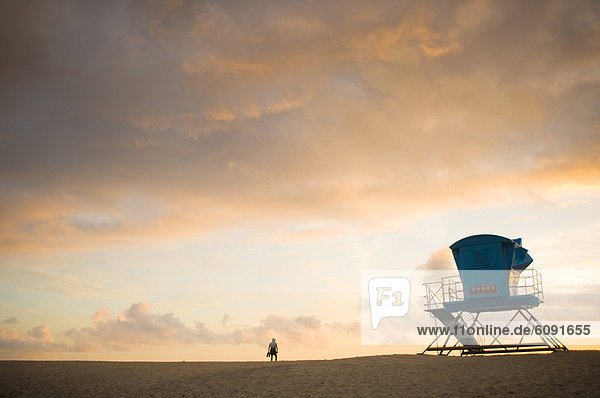 A person next to a life guard tower on the beach watches the sunset.