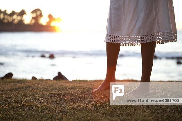 A barefoot woman in a dress walks on grass above the sea at sunset.