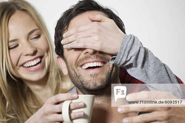 Frau bedeckt die Augen des Mannes und hält Kaffeetasse  lächelnd