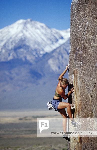 Young woman bouldering in sun with snow capped mountain background.