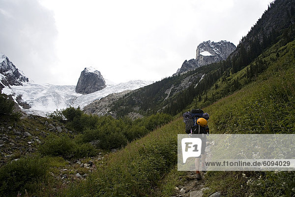 Man carrying a large backpack with a yellow climbing helmet attached to it hikes uphill towards a glacier.