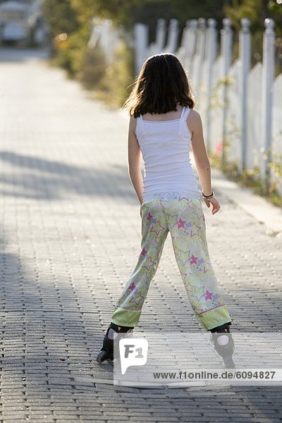 A young girl is roller blading down an alley with a picket fence in the background.