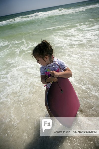 A little girl is holding a body board in the ocean near the shore.