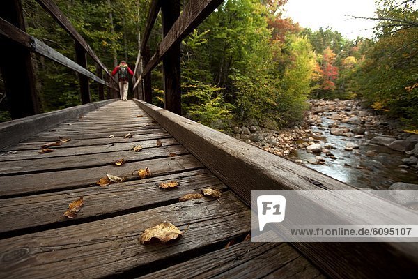 Low angle perspective of one man hiking across a wooden bridge with a stream and fall leaves in view.