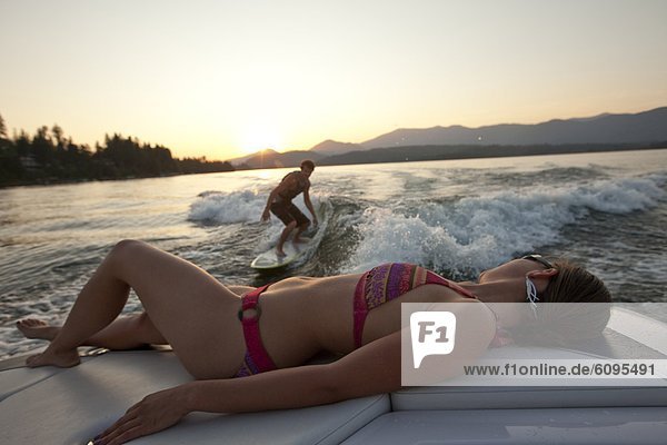 Young man wakesurfing in Idaho as a girl in a bikini watches from the boat.