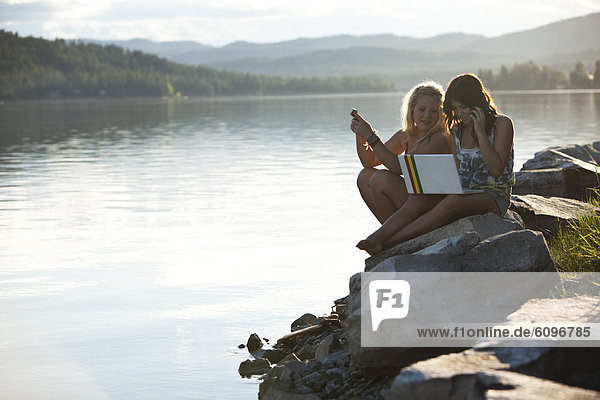 Two young women talk while texting and chatting on a computer on a sunny day at the lake.
