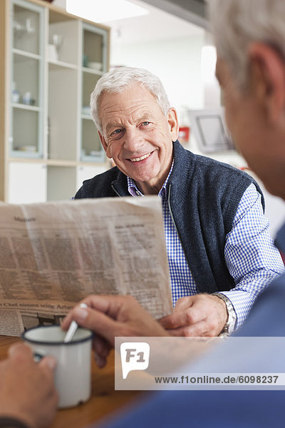 Senior man reading newspaper another man stirring coffee