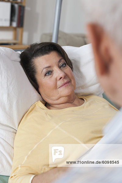 Seniorenfrau auf dem Krankenbett liegend  während der Mann neben ihr sitzt.