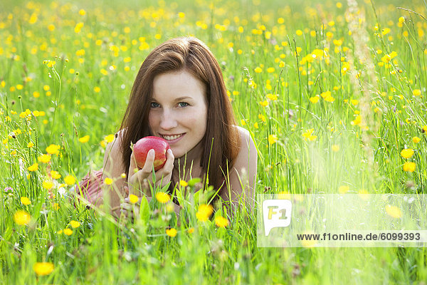 Österreich  Junge Frau im Blumenfeld liegend mit Apfel  lächelnd  Portrait
