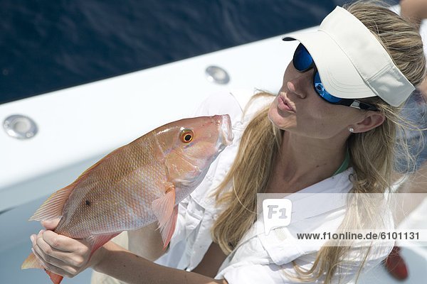 A blonde woman with a white hat and sunglasses holds a freshly caught red snapper and blows it a kiss.