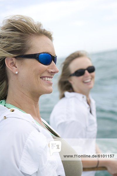 Two women with sunglasses smile with the ocean in the background.