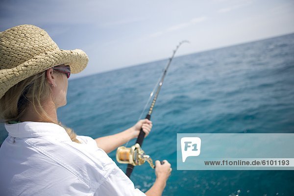 A blonde woman in a straw hat reels in a fish with blue wanter in the distance.