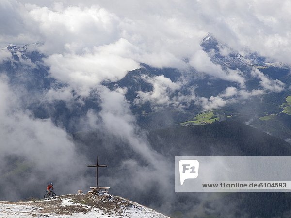 A mountain biker arrives at a cross on the summit of Kronplatz  mountains and clouds in the background.