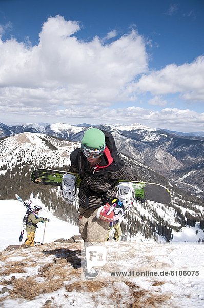 An extreme snowboarder with skiiers in the background climbs to Kachina Peak's summit at Taos Ski Valley  New Mexico.