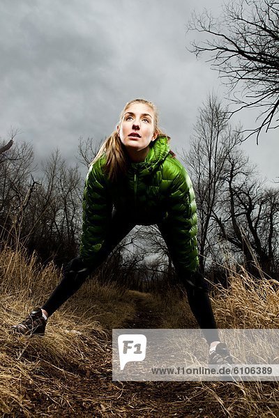 Environmental Portrait of a young woman stretching on a running trail in a green jacket in the winter time.