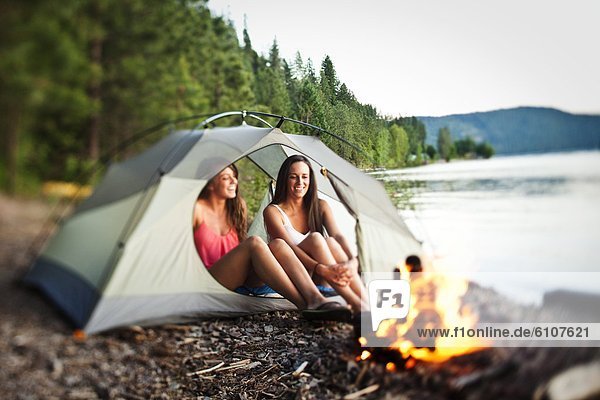 Two beautiful women laugh and smile sitting in their tent next to a campfire in Idaho.