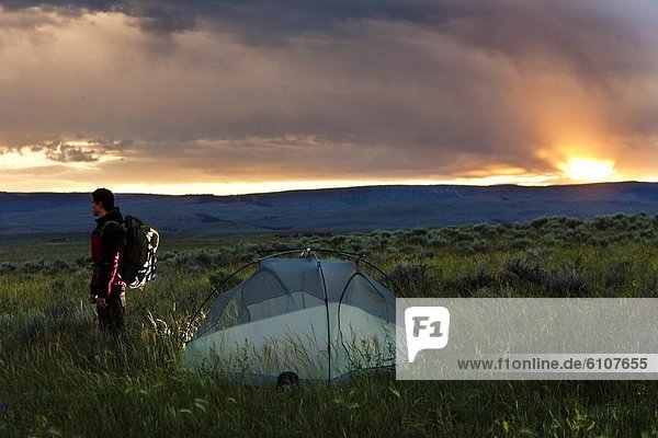 A man standing next to his tent at sunset on a backpacking trip in the mountains of Montana.