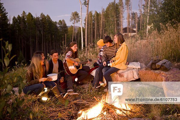 A group of teen-aged girls and boys sing and roast marshmallows around a camp fire on a Summer evening.