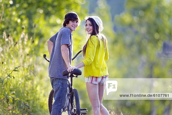 A teen-aged couple and look towards the camera as they walk a cruiser bike down a wooded lane on a Summer afternoon.
