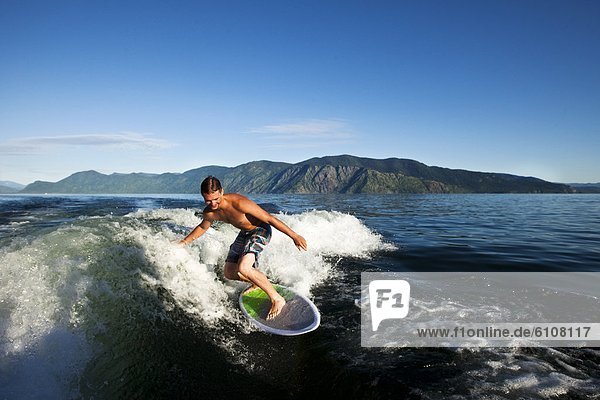 A happy man smiles while wakesurfing behind a wakeboard boat on a sunny day in Idaho.
