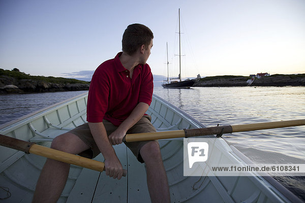 A young man rows in a Peapod dinghy at sunset toward a classic yacht moored along the coast of Maine near Boothbay Harbor.