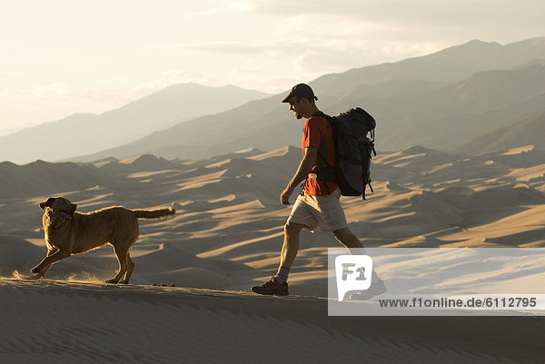 Man hikes with dog in sand dunes.