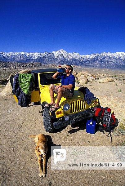 Man sitting on a jeep in the desert with his dog taking a drink from a water bottle.