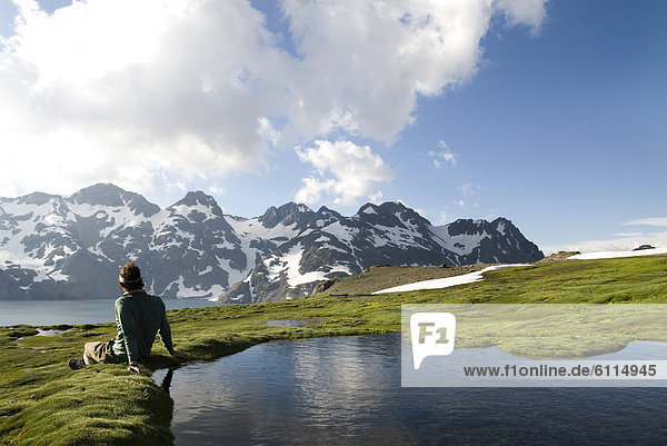 A young man sits next to a small pool enjoying a view of distant snow covered mountains.