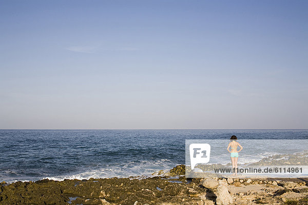 A young woman stands near a small pool on a rocky coast next to the ocean in Jamaica.