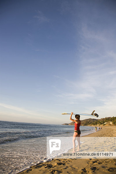 A girl wearing a red top and bikini bottoms holds a surfboard over her head on a remote beach in Sayulita  Mexico.