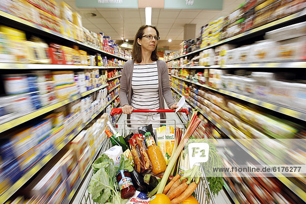 Frau schiebt einen vollen Einkaufswagen durch einen Gang einer Lebensmittelabteilung  Selbstbedienung  Supermarkt  Deutschland  Europa