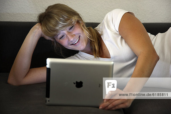 Junge Frau liegt auf einem Sofa und surft mit einem iPad Tablet-Computer drahtlos im Internet