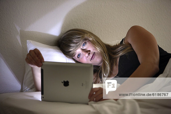 Junge Frau surft im Bett mit einem iPad Tablet-Computer per Drahtlosverbindung im Internet