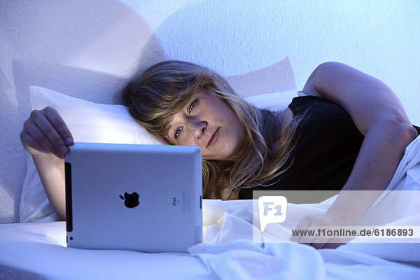 Junge Frau surft im Bett mit einem iPad Tablet-Computer per Drahtlosverbindung im Internet