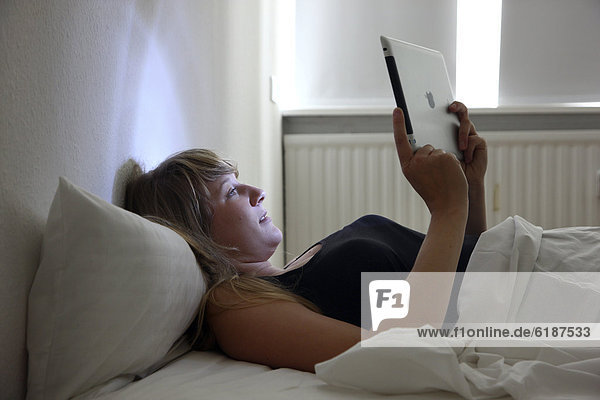 Junge Frau surft im Bett mit einem iPad Tablet-Computer per Drahtlosverbindung im Internet