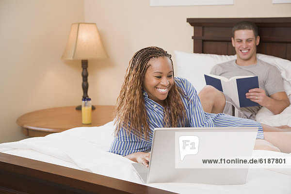 Multi-ethnic couple reading and typing on laptop in bed