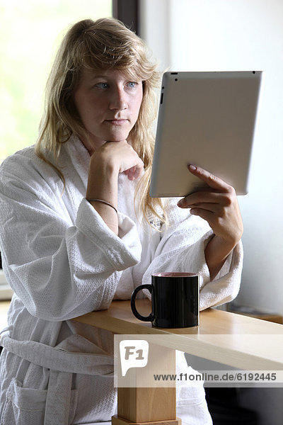 Young woman wearing a dressing gown drinking a cup of coffee in the morning  sitting in the kitchen reading an online newspaper on an iPad  a tablet computer