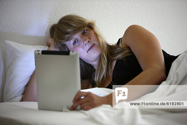 Junge Frau im Bett surft mit einem iPad Tablet-Computer per Drahtlosverbindung im Internet