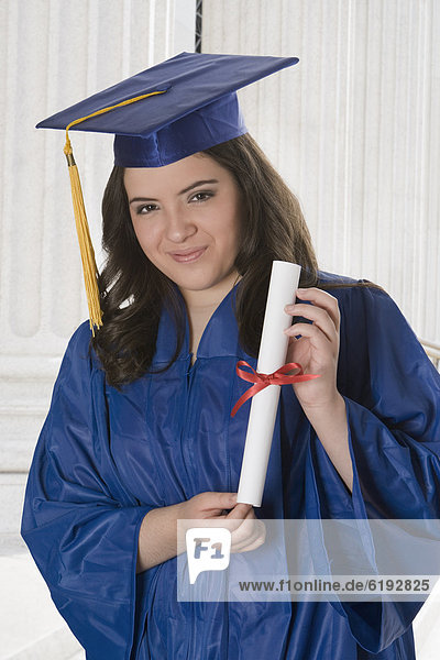 Hispanic teenage girl in graduation cap and gown holding diploma